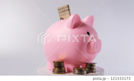Pink piggy bank on a rotating stand with coins and cash around it, white background 121333173