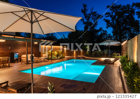Backyard swimming pool with wooden deck and stylish umbrella under evening sky. Modern outdoor lounge area with serene ambiance and elegant lighting 121335427