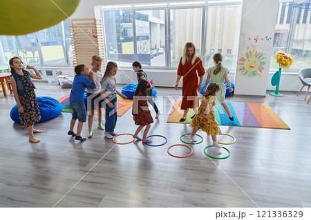 Small nursery school children with female teacher on floor indoors in classroom, doing exercise. Jumping over hula hoop circles track on the floor. 121336329