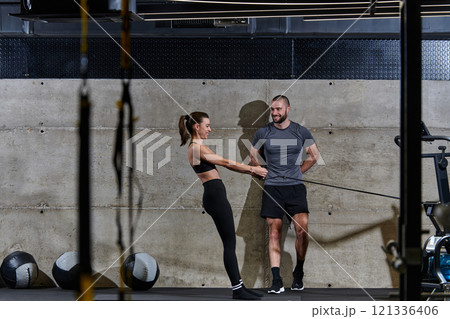 A muscular man assisting a fit woman in a modern gym as they engage in various body exercises and muscle stretches, showcasing their dedication to fitness and benefiting from teamwork and support 121336406