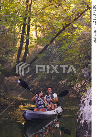 A young couple enjoying an idyllic kayak ride in the middle of a beautiful river surrounded by forest greenery 121337100