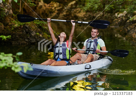 A young couple enjoying an idyllic kayak ride in the middle of a beautiful river surrounded by forest greenery A young couple enjoying an idyllic kayak ride in the middle of a beautiful river surrounded by forest greenery 121337183