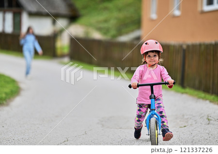 A little girl, dressed in a cute outfit and wearing a helmet, excitedly learning to ride her bike for the first time, filled with joy and determination. A little girl, dressed in a cute outfit and wearing a helmet, excitedly learning to ride her bike for the first time, filled with joy and determination. 121337226