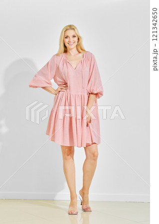 Woman with blonde hair wearing loose pink summer dress and sandals standing over white background 121342650