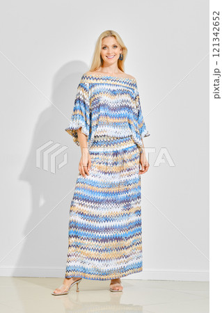Smiling woman in a colourful boho maxi dress standing in bright room over white wall 121342652