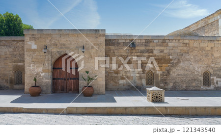 A sunlit courtyard showcases ancient stone walls and a beautifully carved arched wooden door A sunlit courtyard showcases ancient stone walls and a beautifully carved arched wooden door 121343540