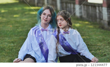 Two female graduates wearing sashes and bow ties are sitting on grass, enjoying their last school day Two female graduates wearing sashes and bow ties are sitting on grass, enjoying their last school day 121344477