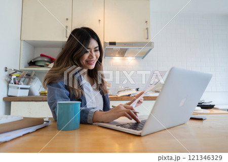 Young Freelance Woman Working from Home in the Kitchen with Laptop and Coffee, Smiling and Reviewing Documents 121346329