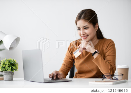 Young Woman Shopping Online and Making a Payment with Credit Card on Laptop in Modern Home Office Setting 121346656