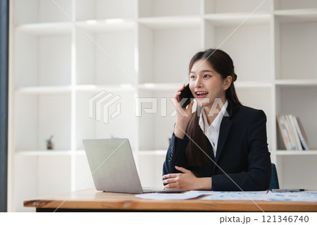 Professional Business Woman Working in Modern Office Environment, Engaged in Phone Call and Using Laptop, Dressed in Formal Attire, Demonstrating Productivity and Professionalism 121346740