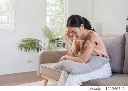 Young Woman Sitting on Sofa Feeling Sick with a Headache, Holding Her Head in Pain, in a Bright and Modern Living Room Young Woman Sitting on Sofa Feeling Sick with a Headache, Holding Her Head in Pain, in a Bright and Modern Living Room 121346742