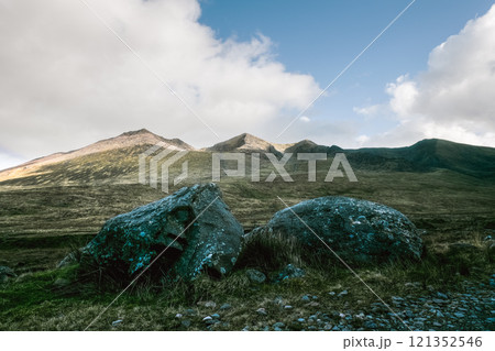 A partly cloudy sky is visible above a range of brown mountains. In the foreground, two large, lichen-covered rocks are present. A partly cloudy sky is visible above a range of brown mountains. In the foreground, two large, lichen-covered rocks are present. 121352546