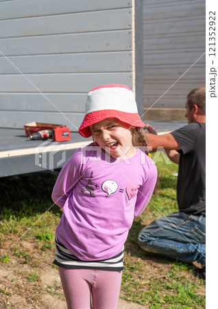 Portrait of a little girl with a funny facial expression against the background of a gazebo being installed. 121352922