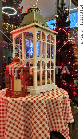 Closeup of cozy Xmas decoration gift box with old vintage lantern form and fir tree illuminated by lights garland on background at outdoors Christmas market at holiday evening or night 121353114