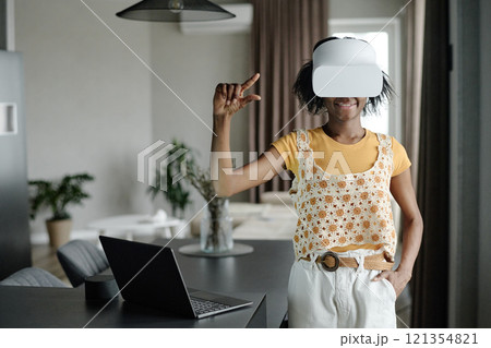 Happy African American woman gesticulating widely and playing VR game while standing in kitchen Happy African American woman gesticulating widely and playing VR game while standing in kitchen 121354821