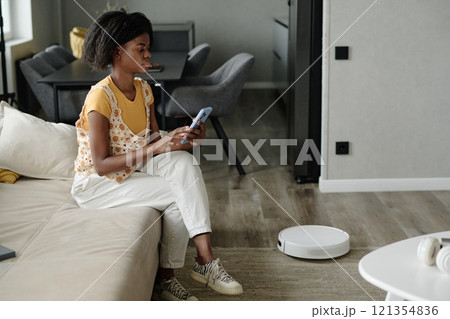 Side view of African American housewife using smartphone app to set robot vacuum cleaner Side view of African American housewife using smartphone app to set robot vacuum cleaner 121354836