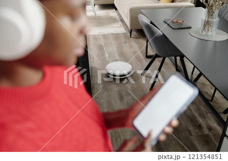 Robot Vacuum cleaning parquet while unrecognizable housewife checking progress on app 121354851