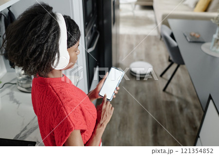 High angle shot of African American woman listening to music while controlling robot vacuum cleaner via smartphone app 121354852