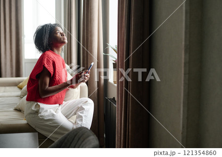 African American woman sitting on couch near huge window while using smartphone in her free time African American woman sitting on couch near huge window while using smartphone in her free time 121354860