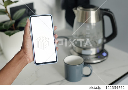High angle hot of smartphone with white screen in unrecognizable female hand, boiling electric glass kettle in background High angle hot of smartphone with white screen in unrecognizable female hand, boiling electric glass kettle in background 121354862