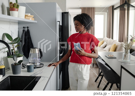 African American woman with smartphone in hand looking at electric glass kettle while waiting for it to boil African American woman with smartphone in hand looking at electric glass kettle while waiting for it to boil 121354865