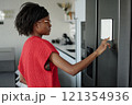 Side view shot of attractive African American woman standing in front of graphite color fridge and setting smart refrigerator 121354936