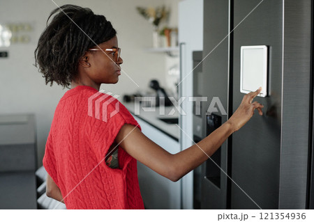 Side view shot of attractive African American woman standing in front of graphite color fridge and setting smart refrigerator Side view shot of attractive African American woman standing in front of graphite color fridge and setting smart refrigerator 121354936