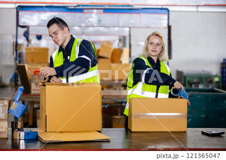 Male and female professional worker wearing safety uniform using packing tape on packaging cardboard box product in warehouse. Male and female professional worker wearing safety uniform using packing tape on packaging cardboard box product in warehouse. 121365047