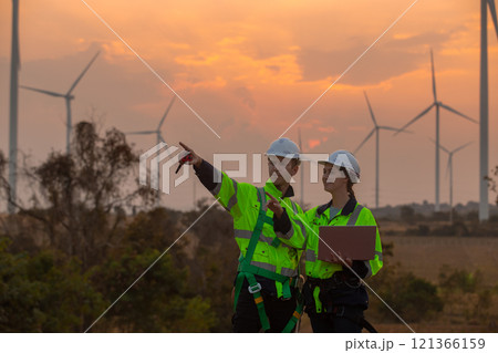 Team engineer wearing safety uniform holding laptop discussed plan about renewable energy at station energy power wind in evening when sunset. Technology environment reduce global warming problems. 121366159