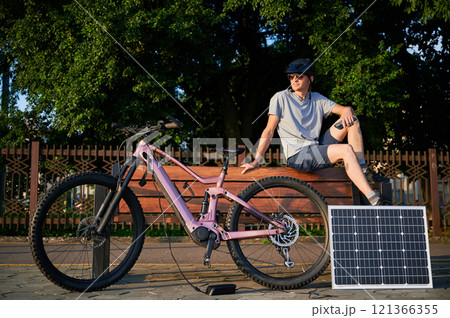 Man cyclist using solar panel for charging electric mountain bike outdoors. Guy in sunglasses sits on bench, holding phone, casually dressed in helmet, t-shirt, shorts, enjoying sunny outdoor setting. Man cyclist using solar panel for charging electric mountain bike outdoors. Guy in sunglasses sits on bench, holding phone, casually dressed in helmet, t-shirt, shorts, enjoying sunny outdoor setting. 121366355