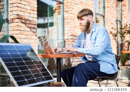 Man works on laptop at outdoor round table, with glass of iced coffee. Solar panel nearby, emphasizing sustainable, modern eco-friendly workspace that combines technology and sustainability. Man works on laptop at outdoor round table, with glass of iced coffee. Solar panel nearby, emphasizing sustainable, modern eco-friendly workspace that combines technology and sustainability. 121366359