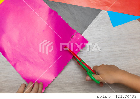 Little child hands holding scissors and cutting colorful geometric shapes from application paper on light blue table background. Pastel color. Making different forms. Point of view shot. Closeup. 121370435