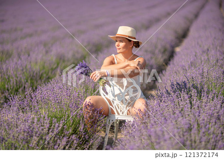 woman sitting in a field of lavender and wearing a straw hat. She is smiling and holding a bouquet of flowers. Scene is peaceful and serene, as the woman is surrounded by the beauty of nature 121372174