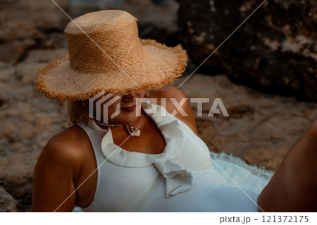 Woman Beach Sunbathing Summer - A woman in a white swimsuit relaxes on a rocky beach while wearing a straw hat, enjoying the summer sun. Woman Beach Sunbathing Summer - A woman in a white swimsuit relaxes on a rocky beach while wearing a straw hat, enjoying the summer sun. 121372175