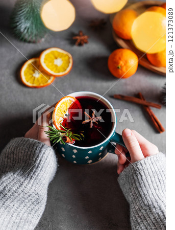 Hands in a sweater holding a cup of mulled wine with orange, cinnamon and rosemary  121373089