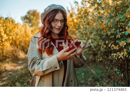 Autumnal style trendy shot. Stylish woman in hat and style clothes with selfie on smartphone at park with orange leaves Autumnal style trendy shot. Stylish woman in hat and style clothes with selfie on smartphone at park with orange leaves 121374865