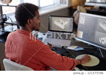 African American man focusing on creating architectural designs on large screen in modern office setting. Multiple digital devices and papers on desk supporting detailed work environment 121375596