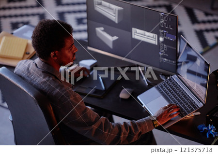 African American man working with virtual 3D models on dual computer screens in dark room with focused lighting. Using modern gadgets and design software with attention to detail 121375718