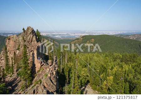A group of tourists sits on an igneous rock against the backdrop of a green forest and a city in the distance A group of tourists sits on an igneous rock against the backdrop of a green forest and a city in the distance 121375817