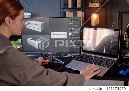 Woman working at desk with dual monitors and laptop, focusing on 3D modeling of furniture. Screens displaying detailed 3D designs of couch 121375970