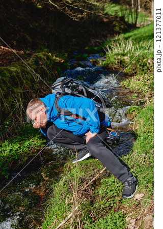 Hiker drinking stream water in mountain. Adult man drinking water from the mountain creek. Tourist wearing casual clothes and sunglasses making a sip of mountain river water from the palms of his 121377011