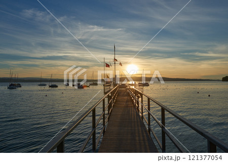 A serene sunset over a calm river, with vibrant orange and pink hues in the sky. A small sailboat is anchored in the water, reflecting the colorful sky. 121377054