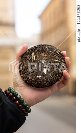 A hand is holding a unique compressed tea cake against an enchanting city landscape backdrop 121378362