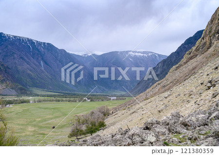 View from the top of a deep canyon surrounded by mountains, a river flowing below and a steep winding road descending down to the river on an overcast spring day. Chulyshman river valley, Altai, 121380359