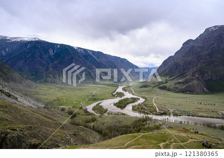 View from the top of a deep canyon surrounded by mountains, a river flowing below and a steep winding road descending down to the river on an overcast spring day. Chulyshman river valley, Altai, 121380365
