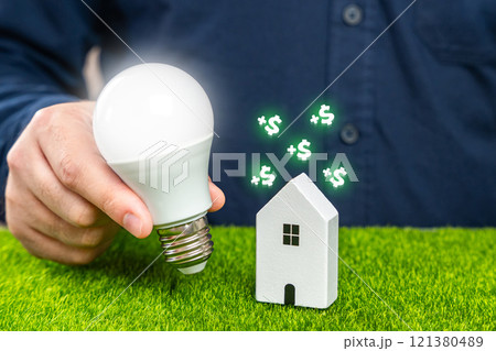 Save electricity at home and gain money. Utilities services. Save on bills. A man holds a glowing light bulb near a house. Save electricity at home and gain money. Utilities services. Save on bills. A man holds a glowing light bulb near a house. 121380489