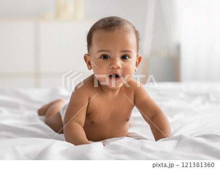 Portrait of cute small African American baby crawling on bed on the white blanket at home. Naked black infant lying on belly, looking at camera. Free copy space, selective focus, blurred background Portrait of cute small African American baby crawling on bed on the white blanket at home. Naked black infant lying on belly, looking at camera. Free copy space, selective focus, blurred background 121381360