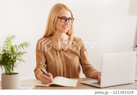 A woman sits at a desk, working on her laptop while taking notes in a notebook. She is smiling and looking focused. 121381429