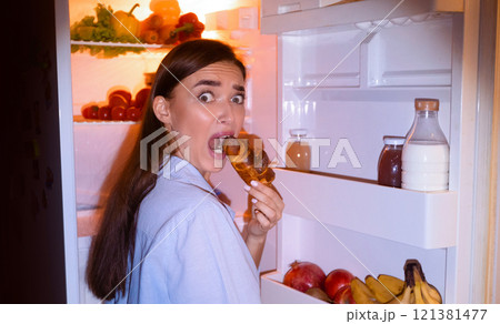 Midnight snack. Scared girl in pajamas eating croissant near opened fridge, free space Midnight snack. Scared girl in pajamas eating croissant near opened fridge, free space 121381477