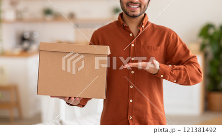 Cropped of smiling man in a red shirt holds a cardboard box in front of him, his other hand extended towards the viewer. The background is blurred, creating a soft and inviting atmosphere. Cropped of smiling man in a red shirt holds a cardboard box in front of him, his other hand extended towards the viewer. The background is blurred, creating a soft and inviting atmosphere. 121381492